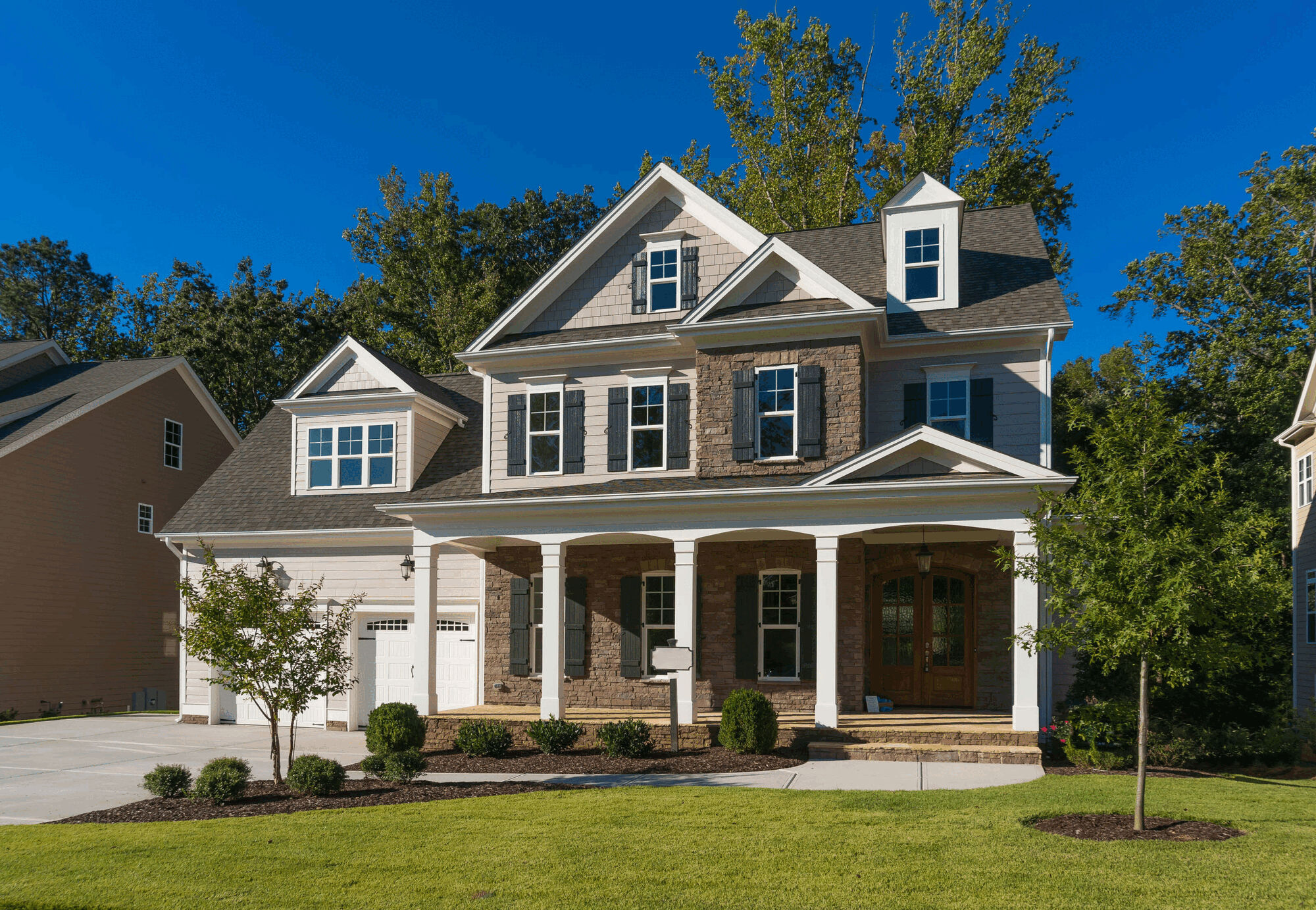 Two-story suburban house with front porch, garage, and landscaped lawn under clear blue sky.