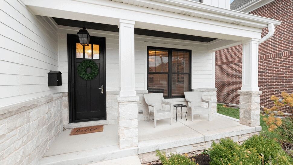 Front porch with black entry door, white siding, stone columns, and two chairs