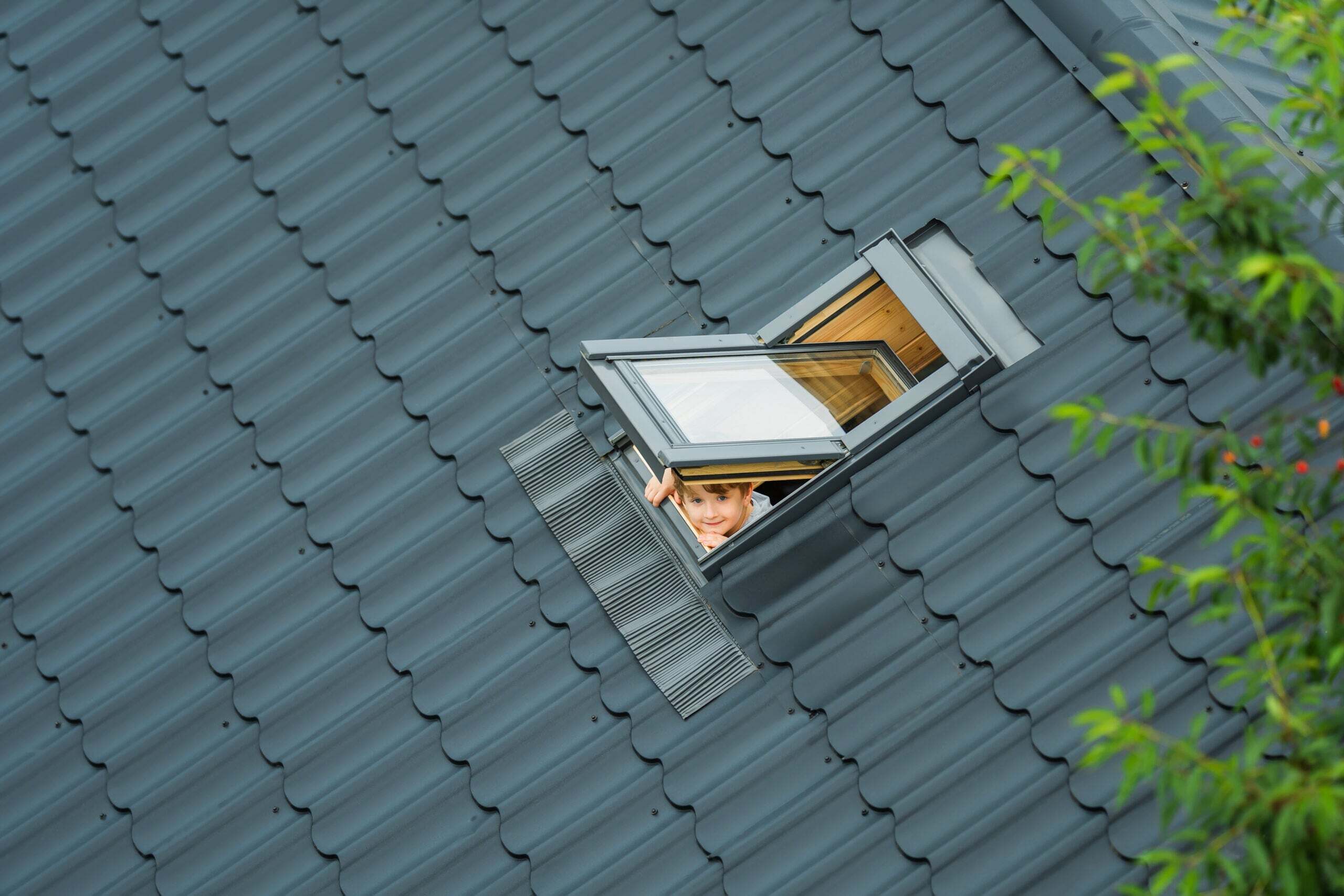 Little cheerful boy looks up into an open skylight window on roof of house covered with metallic gray tiles. Child breathes clean air. Summer morning. Top view.