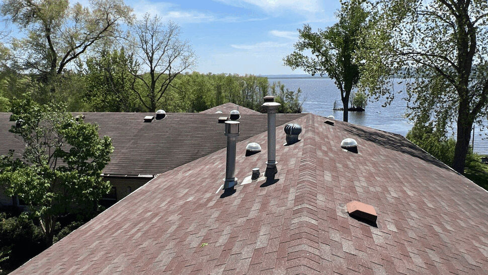 Asphalt roof inspection by the lake | First American Roofing & Siding Inc Asphalt shingle roof ridge with vents overlooking a lake and treeline.