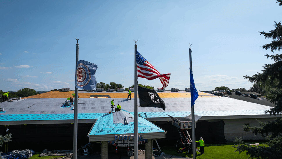 Roofer crew installing blue underlayment on a large roof with flags.