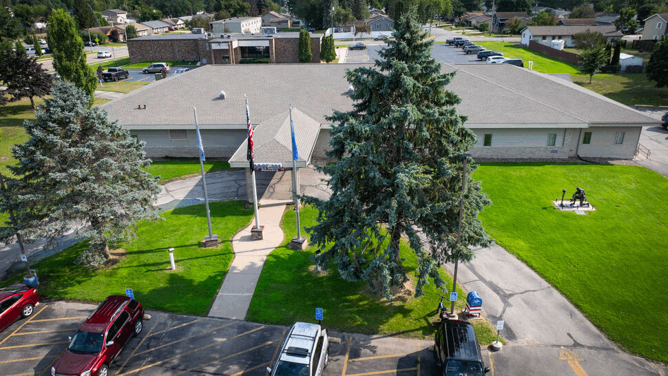 Front Entrance Roof Aerial | First American Roofing & Siding Inc Front entrance, flagpoles, and roof framed by large evergreen trees.