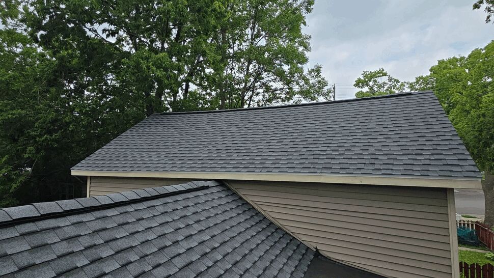 Intersection of two gray asphalt shingle roofs with trees behind.