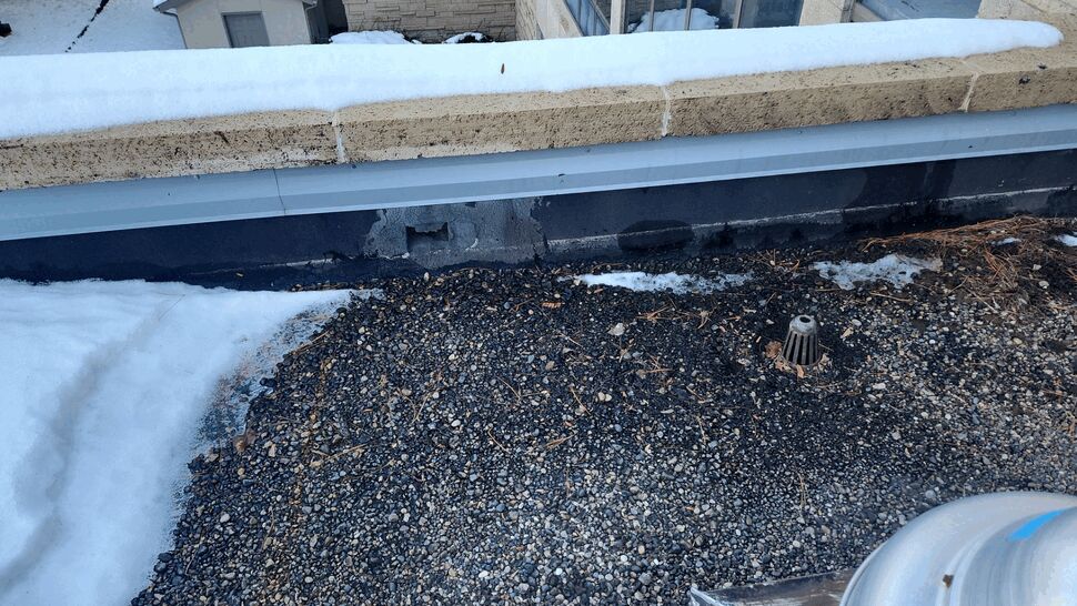 Winter Roof Flashing Damage | First American Roofing & Siding Inc Snow-covered commercial roof edge showing damaged wall flashing and gravel surfacing