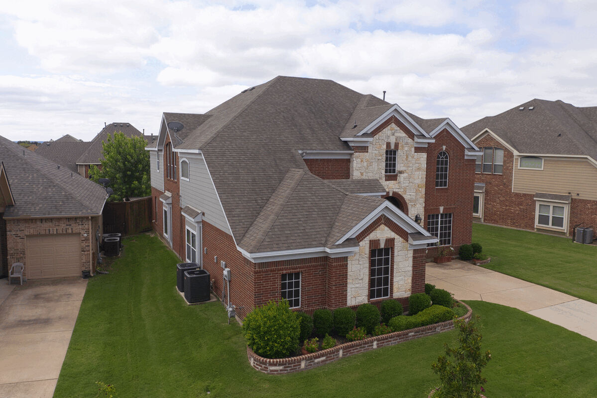 Aerial side view of a multi-gabled brick house roof and surrounding lawns.
