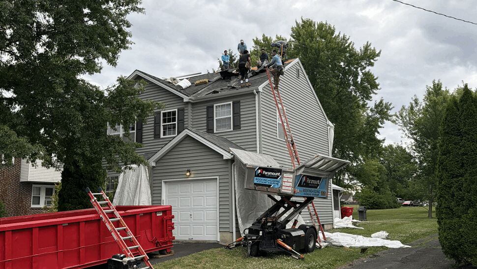 Workers on a two-story home's roof installing shingles with dumpster and lift equipment.