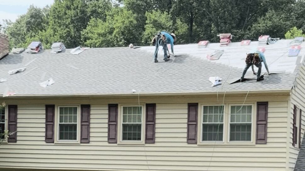 Two roofers installing shingles on a house roof secured with safety ropes.