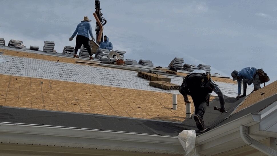 Roofers working on an exposed roof deck laying underlayment with stacked shingles.