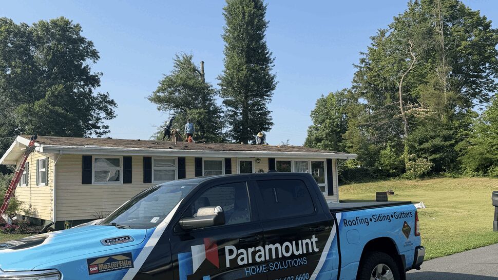 Blue Paramount Home Solutions pickup parked with crew working on a house roof under clear sky.