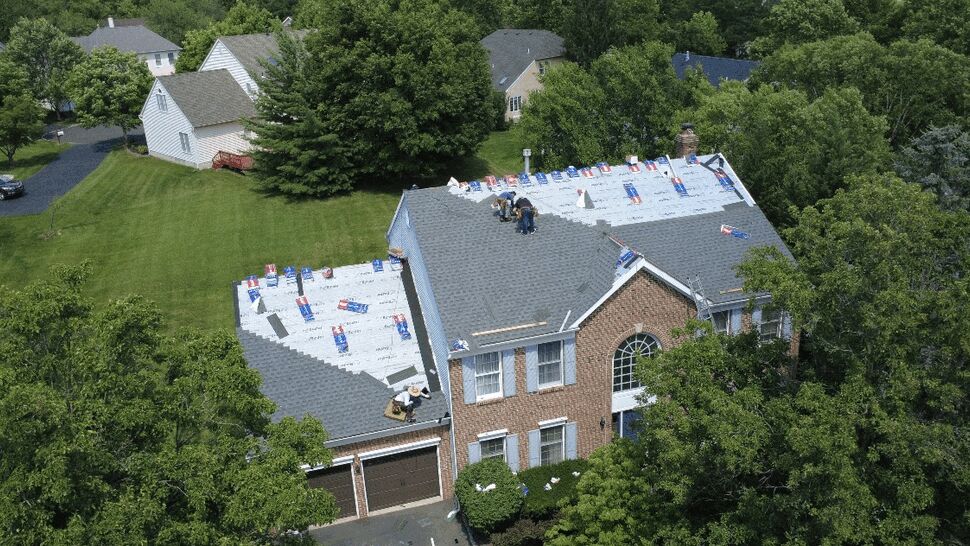 Drone view of workers installing shingles on the front-facing roof of a brick two-story home.