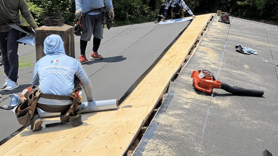 Workers installing plywood and rolling out underlayment across a roof deck.