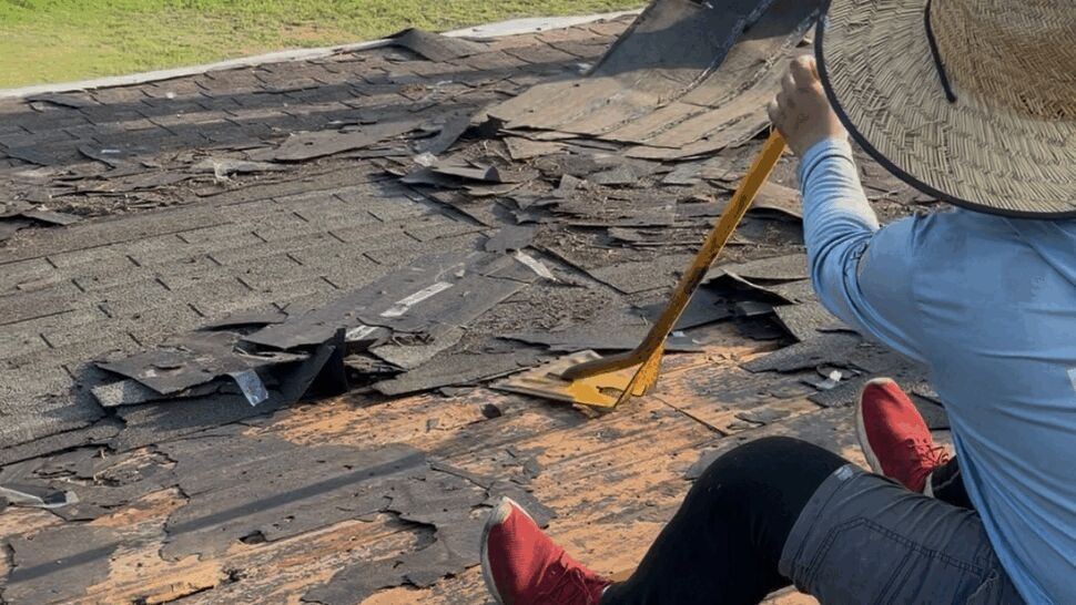 Workers tearing off old shingles from a roof with tools and debris visible.