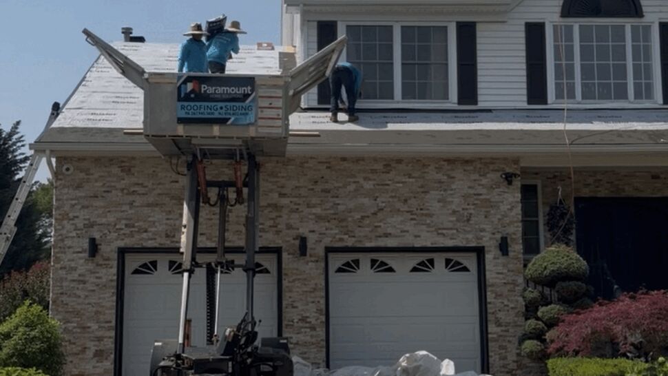 Workers on a material lift installing roofing above a two-car garage; Paramount sign visible on equipment.