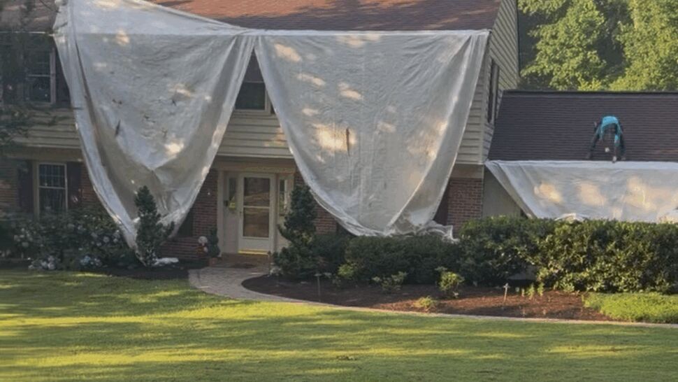 Two roofers on a roof with large protective tarps draped over the front of a house.