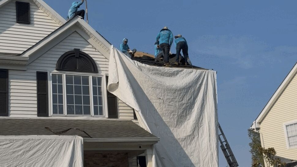Crew of roofers working on the upper roof with a white tarp draped down the side of the house.