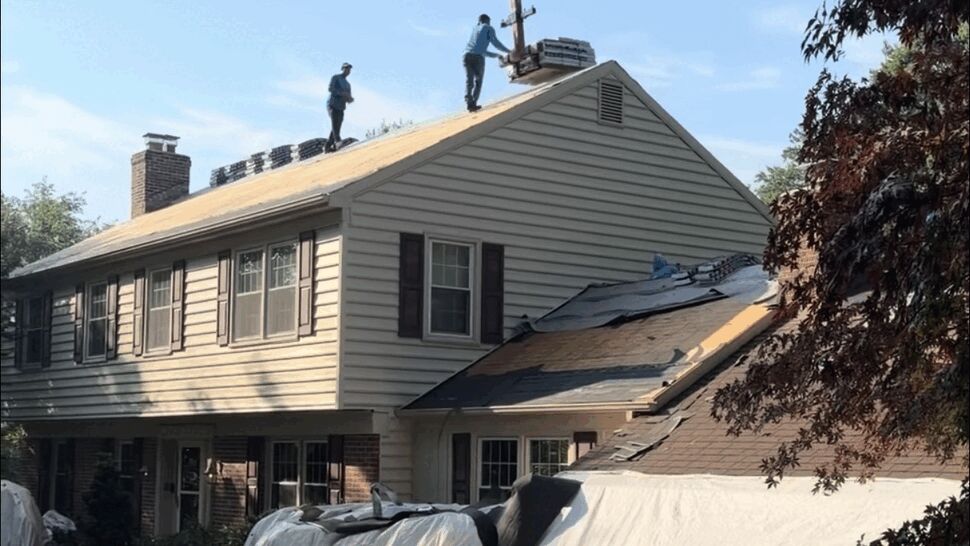 Two workers on a sloped roof unloading bundles of shingles from a crane.