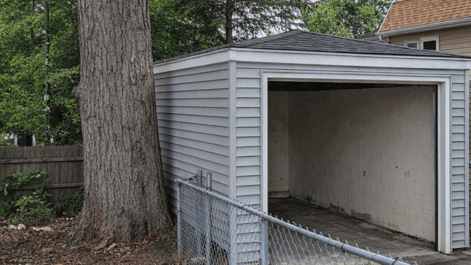 Open Vinyl Garage with Siding | Tycos Roofing And Siding Inc Small gray vinyl-sided garage with open bay door beside a large tree