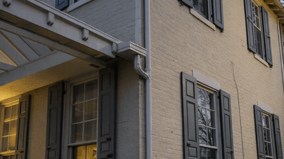 Evening view of a yellow brick house corner with gray shutters and downspout