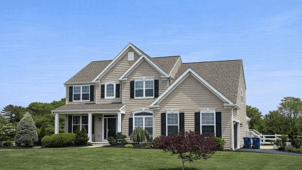 Two-story beige house with porch, black shutters, and landscaped lawn.