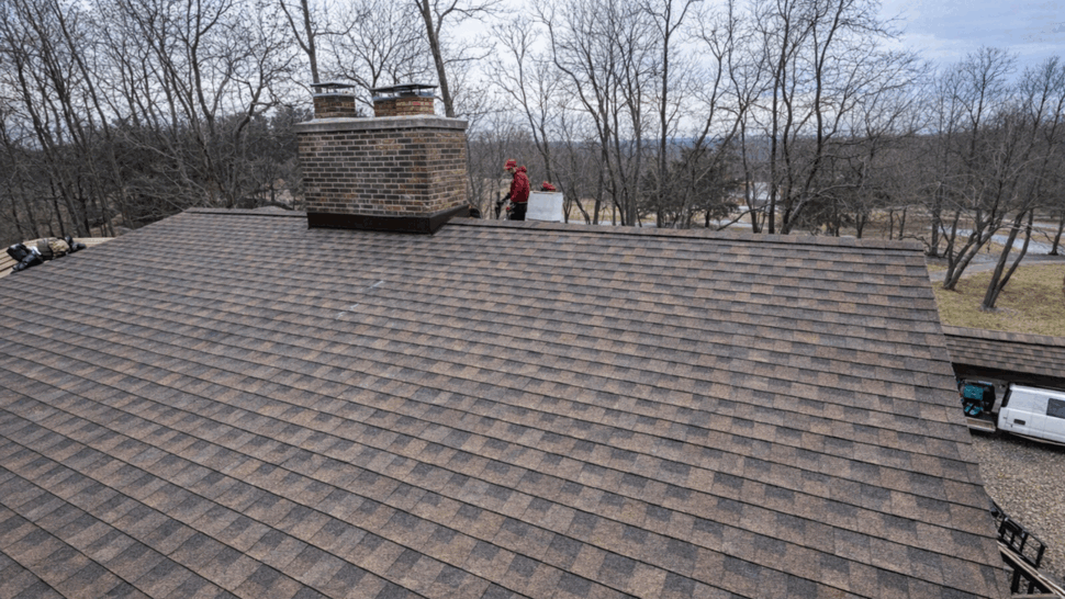 Crew installing brown shingles near brick chimney on residential roof