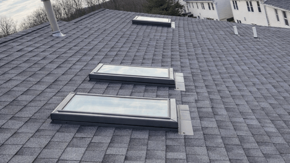 Three skylights on shingled roof | Tycos Roofing And Siding Inc Three rectangular skylights installed on a gray asphalt shingle roof with trees in the distance.