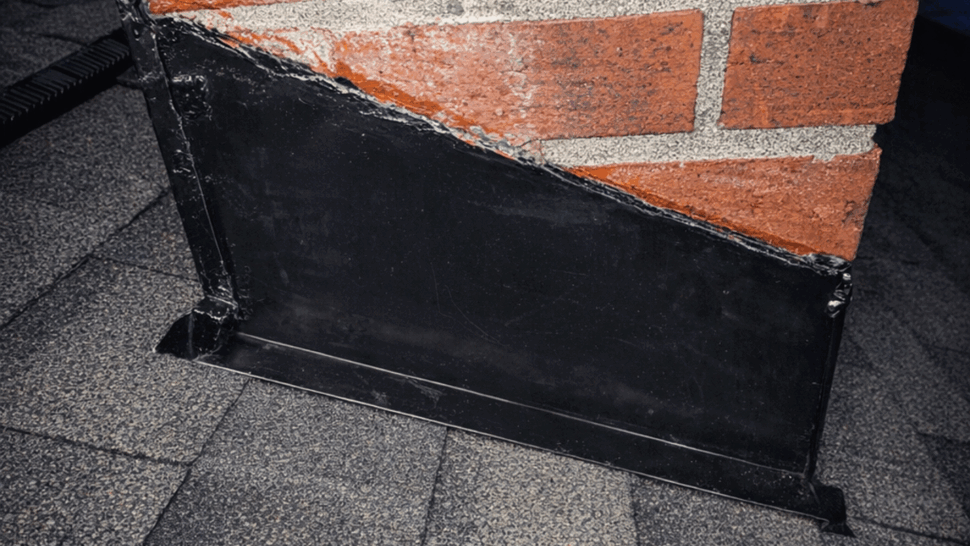 Chimney flashing repair on shingled roof | Tycos Roofing And Siding Inc Close-up of black metal chimney flashing and sealant against a brick chimney on asphalt shingles.