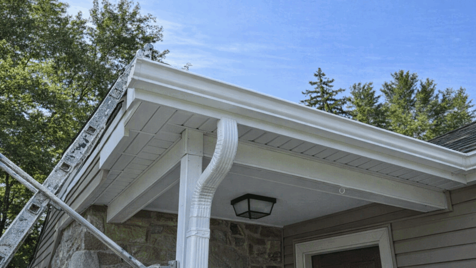 Gutter and downspout detail at porch | Tycos Roofing And Siding Inc Close-up of white gutter and curved downspout at a home's porch soffit with a ladder nearby