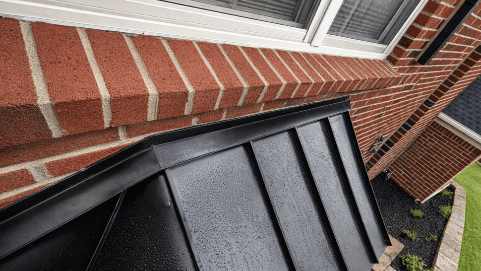 Close-up of black standing-seam metal roof panels with water droplets next to a brick window sill.