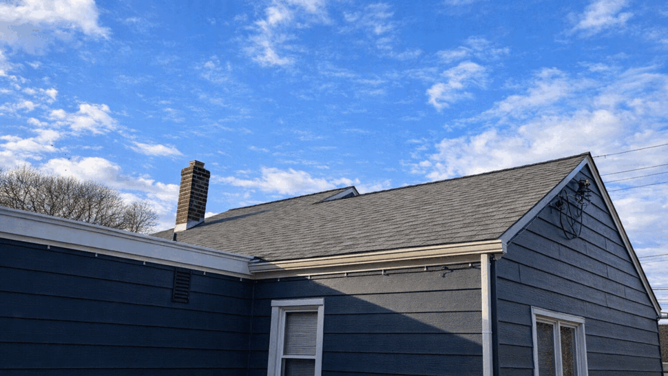 Gable Roof and Gutter Detail | Tycos Roofing And Siding Inc Close view of a gray shingle roof, gutter, chimney, and blue siding.