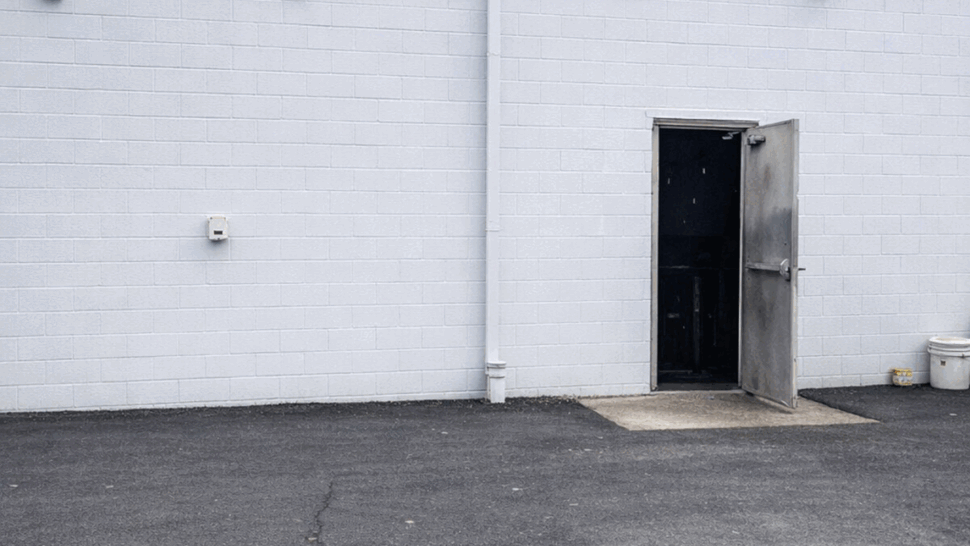 Open metal service door in a white cinderblock wall with downpipe and yellow gas line above.