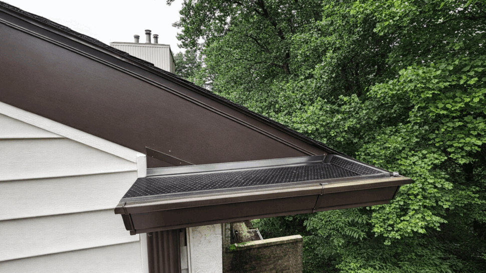 Brown gutter with gutter guard along a sloped roof edge and trees.