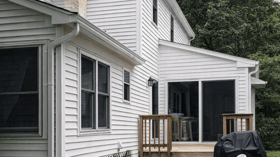 Backyard view of a small wooden deck, sliding door, patio and white vinyl siding.