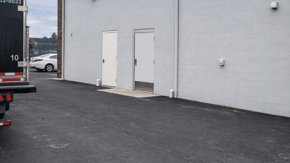 Side of a white commercial building with two doors, parked truck, and freshly paved asphalt under cloudy sky.