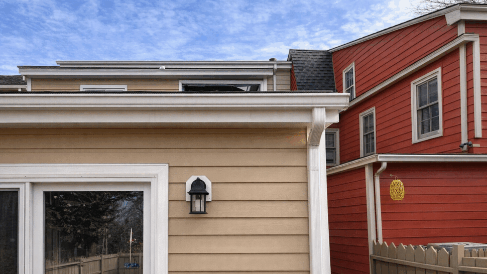 White gutter corner and downspout attached to beige siding between houses.