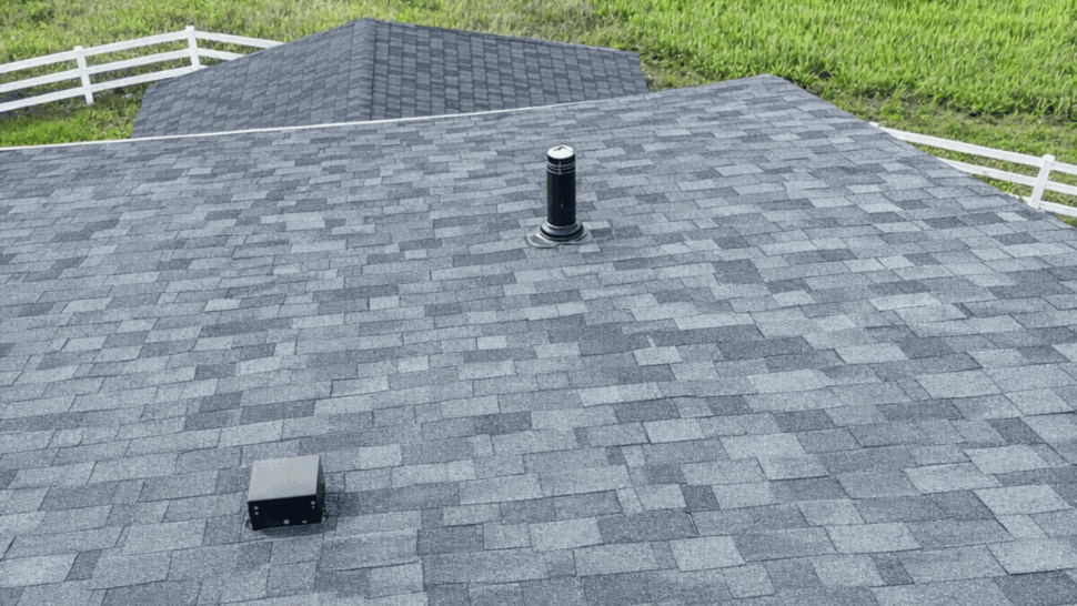 Asphalt Shingle Close-Up with Roof Vent | Tycos Roofing And Siding Inc Close-up of dark gray asphalt shingle roof with a plumbing vent and green field beyond.