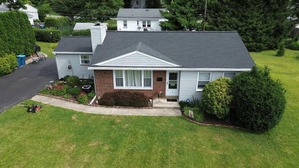 Aerial front view of a small ranch house with dark asphalt shingle roof and green lawn.