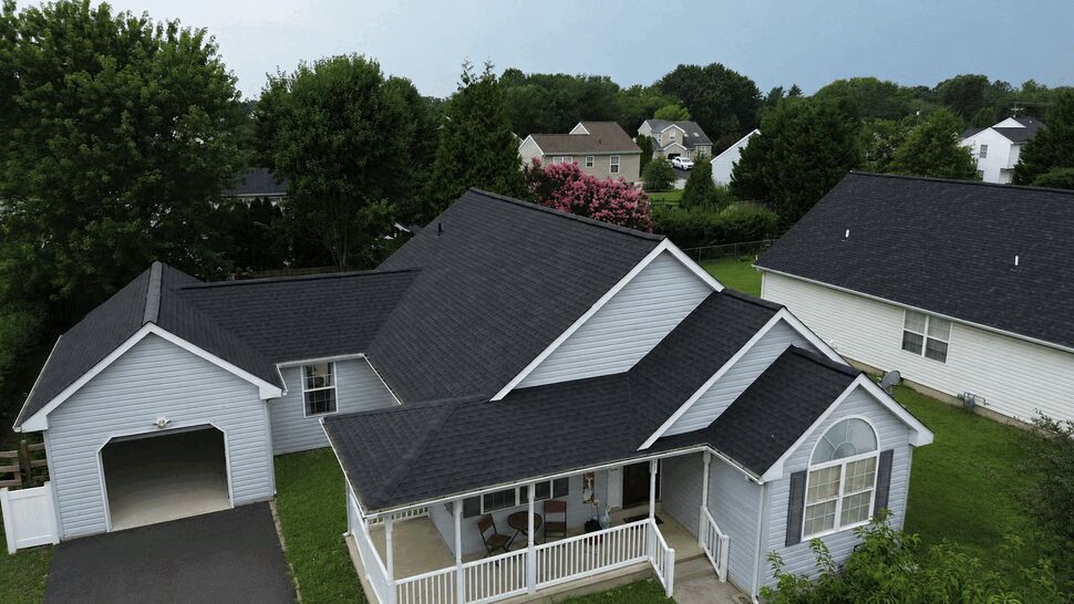 Aerial view of a light-gray house with black shingle roof and front porch.