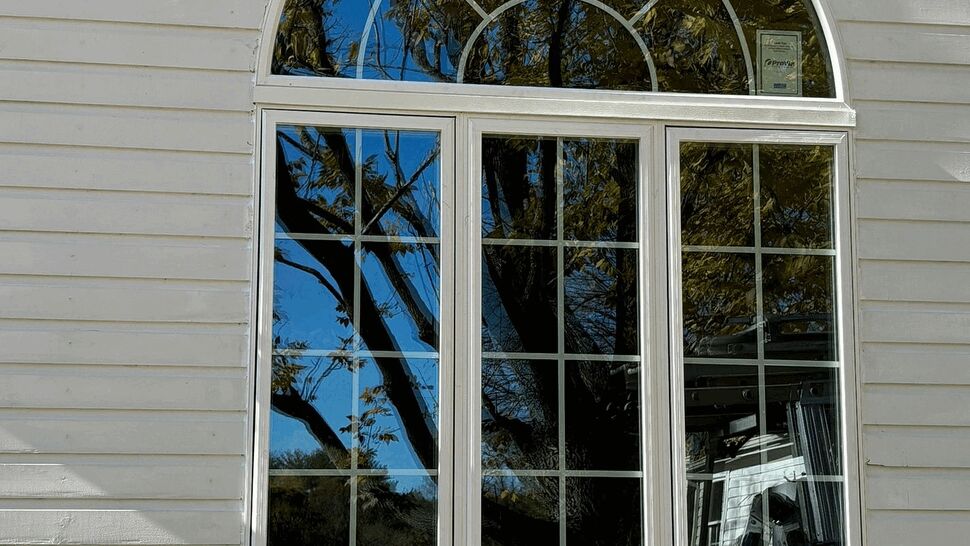Arched window with grid muntins reflecting trees on white siding