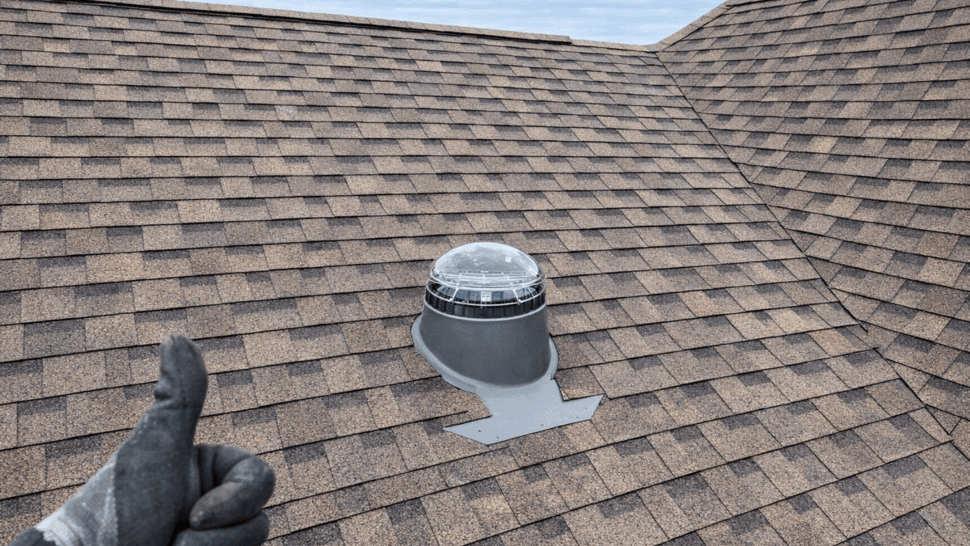 Close-up of brown asphalt shingle roof with installed tubular skylight and gloved thumbs-up