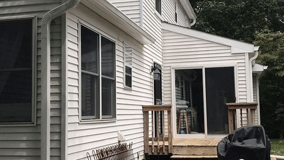 Backyard view of a small wooden deck, sliding door, patio and white vinyl siding.