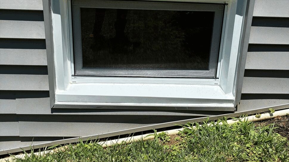 Small basement window with white trim set in gray vinyl siding
