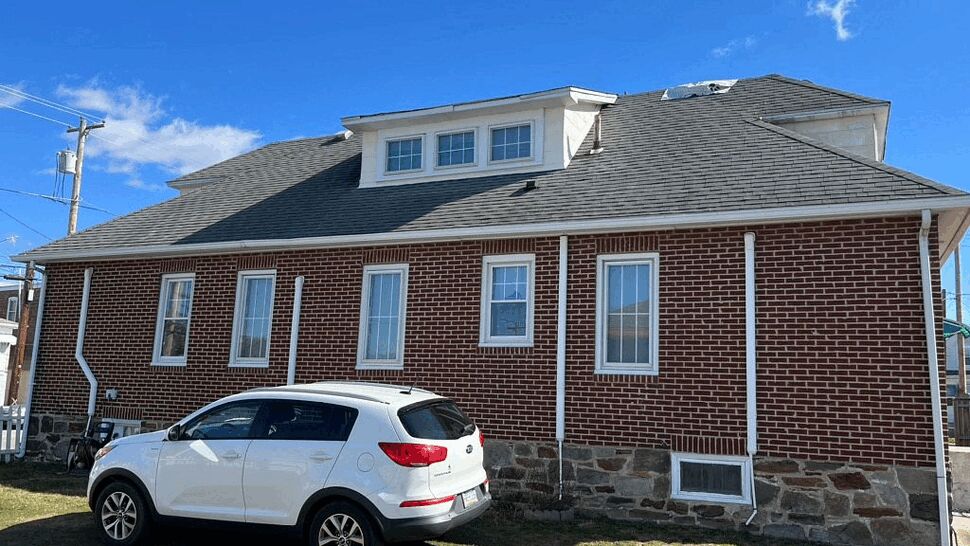Side of a brick house showing multiple windows, downspouts, and a white SUV parked on the lawn.