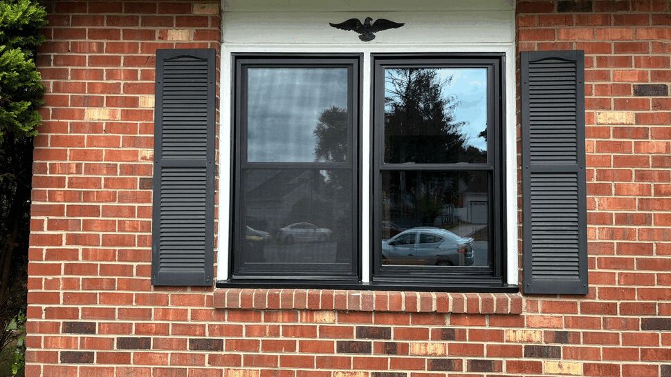 Brick Wall Window Upgrade | Tycos Roofing And Siding Inc Black-framed replacement window with shutters set in a brick wall.