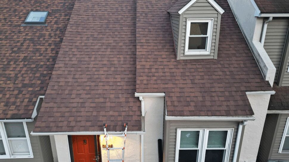 Aerial view of brown shingle roof with dormer and ladder over red entry door