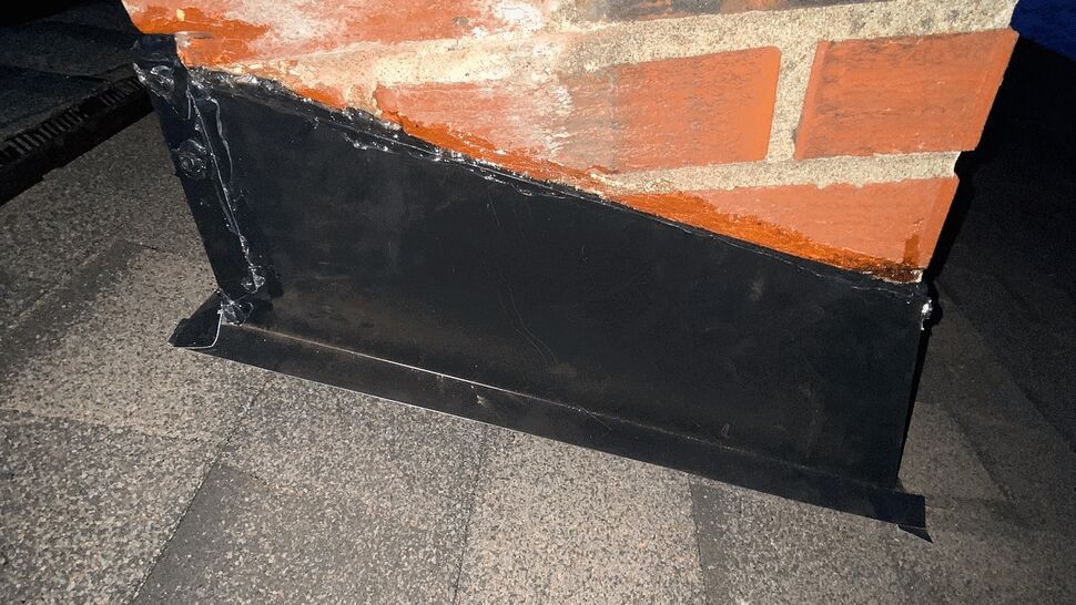 Close-up of black metal chimney flashing and sealant against a brick chimney on asphalt shingles.