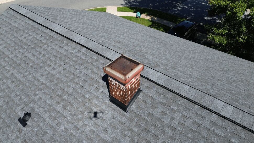 Chimney and ridge vent on new roof | Tycos Roofing And Siding Inc Aerial shot of a brick chimney and ridge vent on a gray shingle roof.