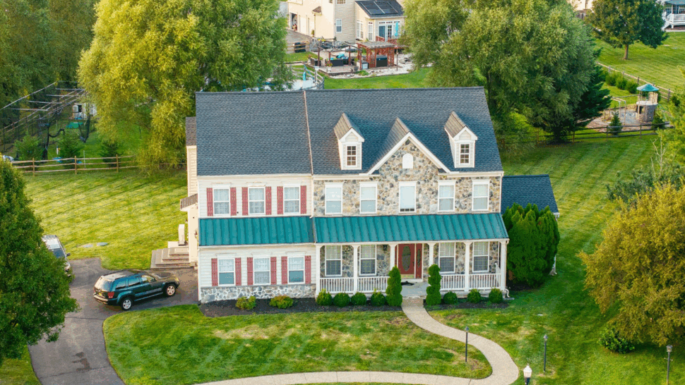 Aerial front view of colonial home with dark shingle roof and green porch awning