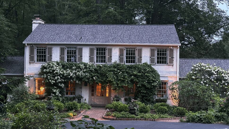Two-story colonial home with shingle roof, shutters, and illuminated windows at dusk