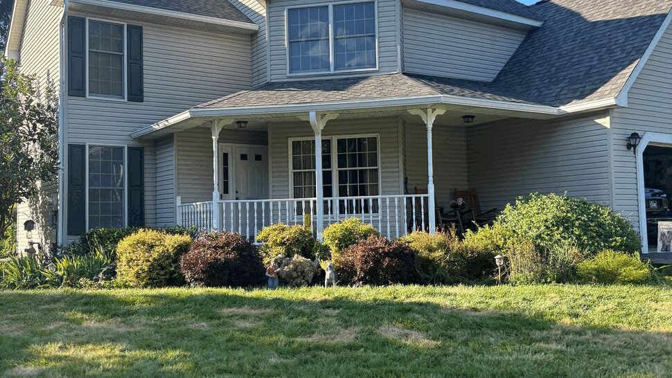 Two-story beige house with front porch, vinyl siding and dark shingle roof under blue sky.