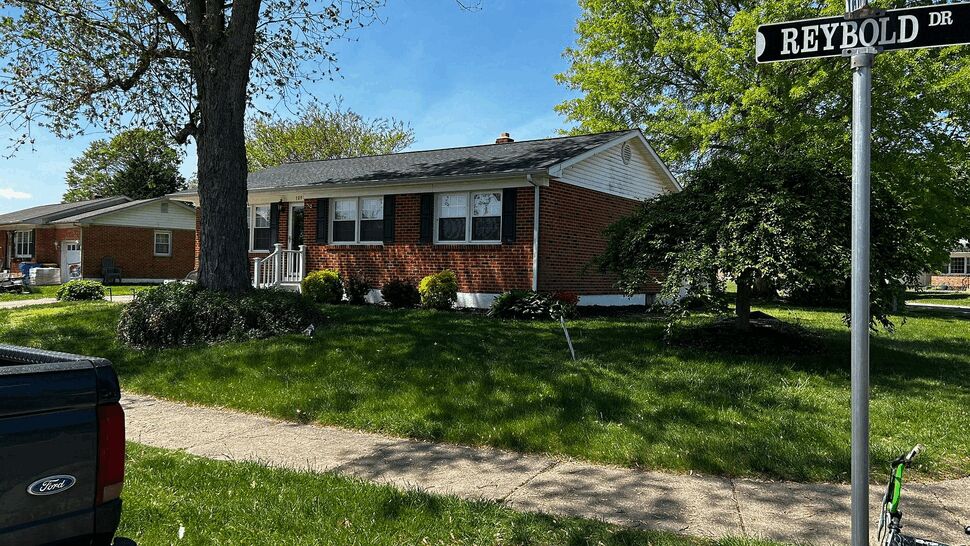 Corner view of brick home with tree, sidewalk, and shingle roof.